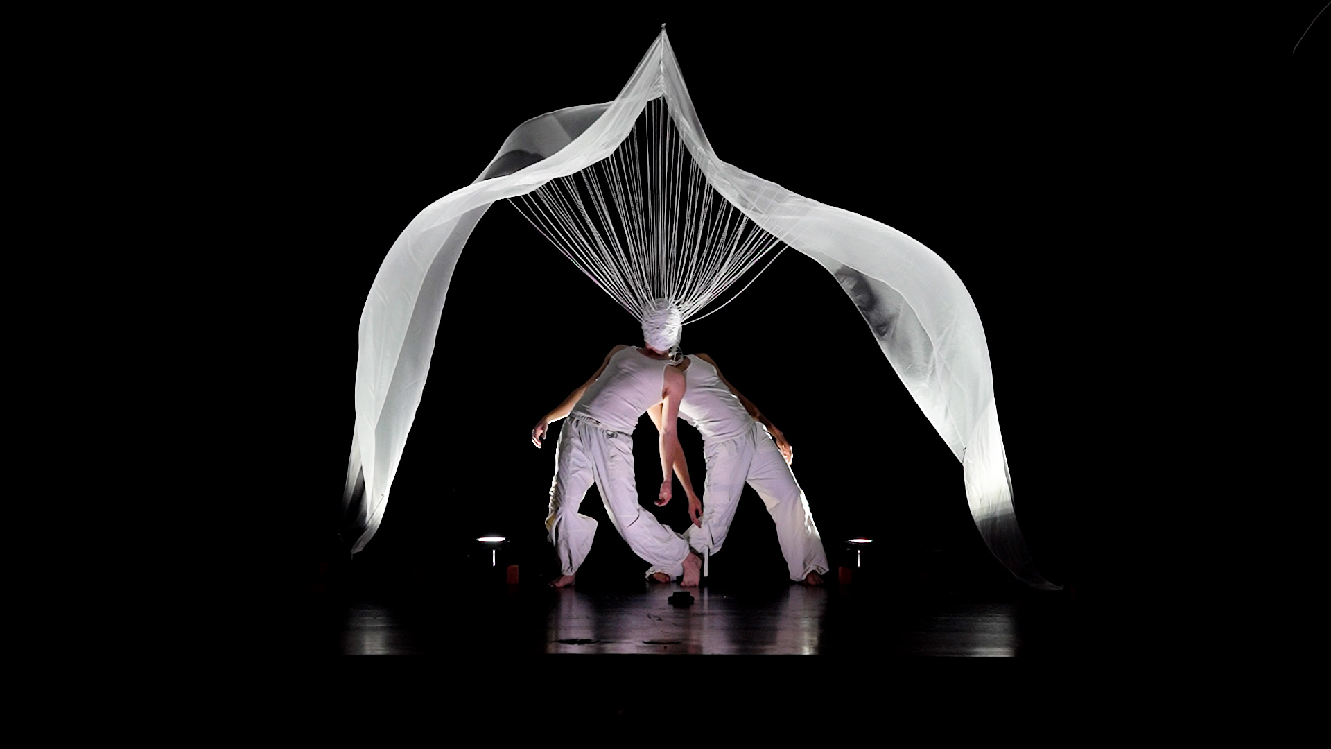 two figures in white standing back-to-back beneath stretched fabric tied to a head-knot suspended above